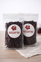 Deux sachets de bissap séché présentés sur une table en bois, à côté d’un tissu clair.